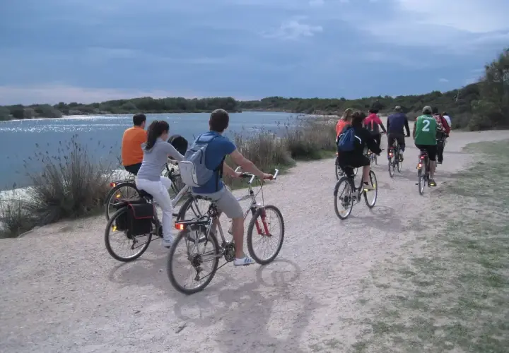 Grupo de ciclistas pedaleando por un camino de tierra junto a la albufera de Valencia. El cielo está nublado y el agua refleja la luz.