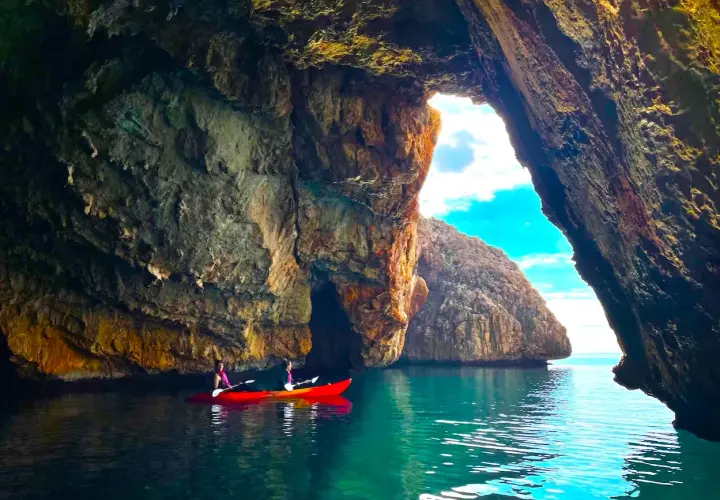 Dos personas en kayak rojo exploran la cueva de la cala del llop mari, con aguas turquesas y formaciones rocosas impresionantes.