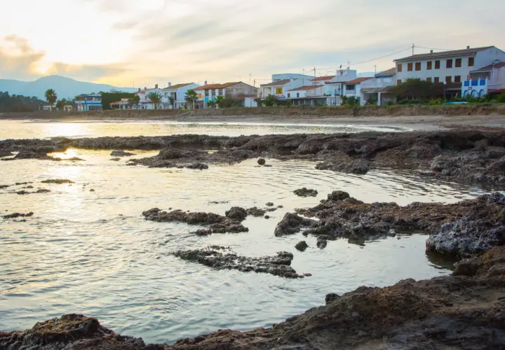 Costa con casas blancas y palmeras, rocas volcánicas en primer plano y el mar reflejando la luz del atardecer. Se divisa una torre la sal a lo lejos.