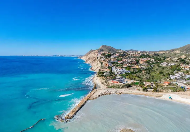 Vista aérea de la costa con casas blancas y el mar azul turquesa. La cala Baeza se extiende hacia el horizonte, con una bahía protegida por un rompeolas de rocas.
