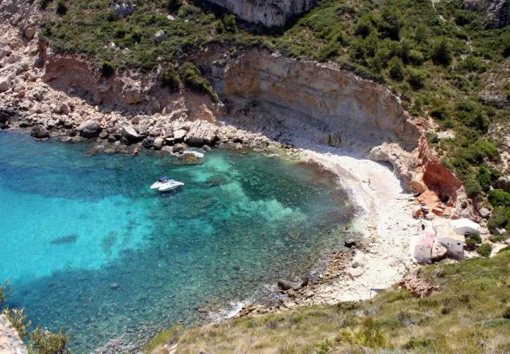 Aguas turquesas y una cala rocosa en Cala Llebeig, con un pequeño barco y casitas blancas en la orilla.
