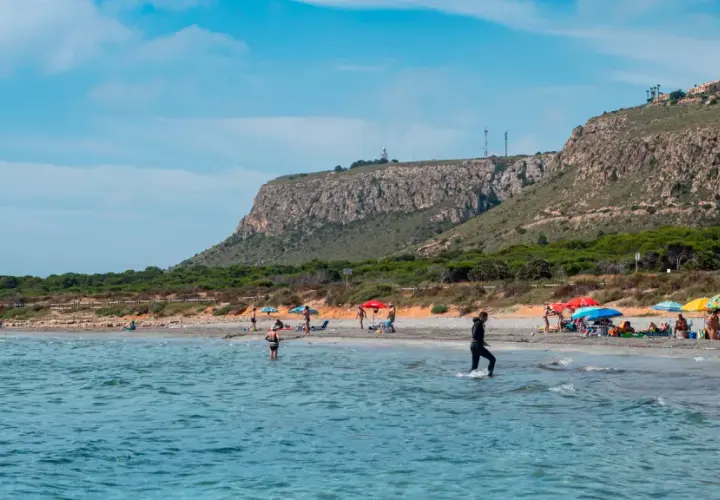 Gente disfruta del sol en la playa de la ermita santa pola, con aguas turquesas y un acantilado verde al fondo.