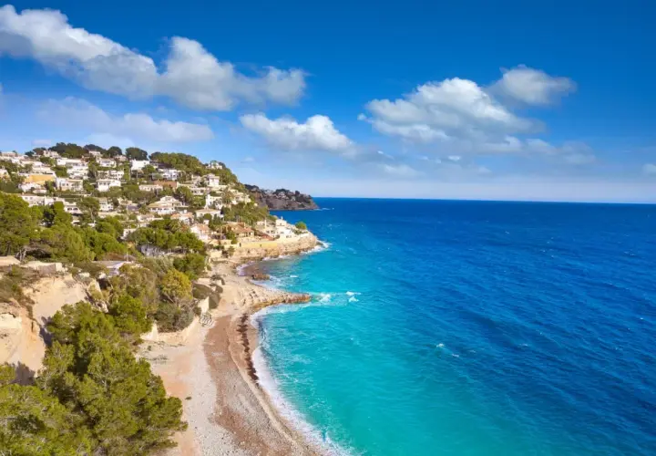 La **cala Baladrar** se asoma entre casas blancas y pinos verdes, con aguas turquesas y un mar azul intenso bajo un cielo nublado.