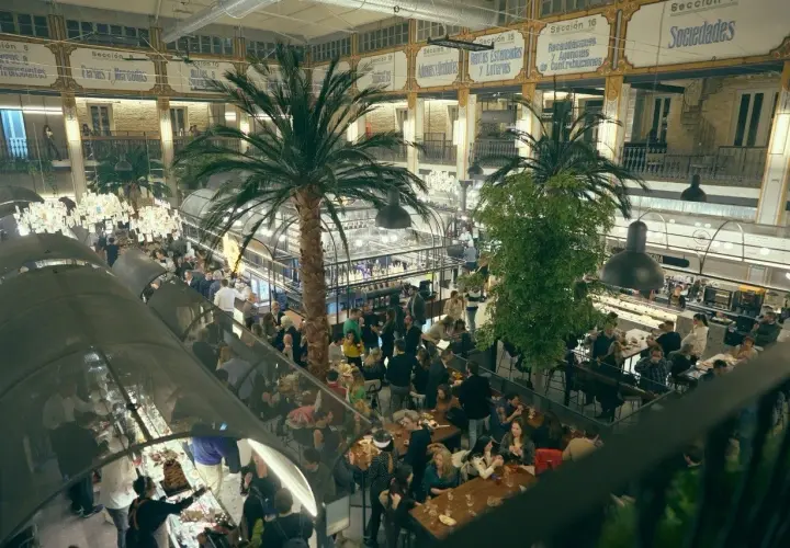 Un bullicioso mercado interior con palmeras, gente comiendo y comprando. El ambiente recuerda a un mercado de la imprenta, lleno de vida y actividad.