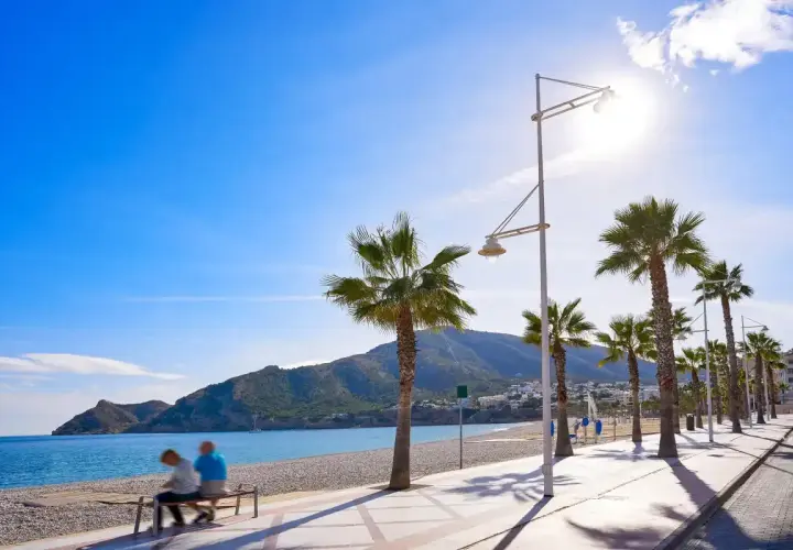 Pareja sentada en un banco en la playa cap blanc, con palmeras y montañas al fondo bajo un cielo azul brillante.