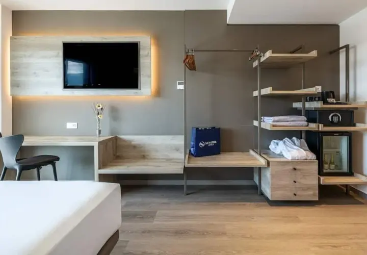 Habitación moderna con TV, escritorio, armario abierto con minibar y caja fuerte. Una bolsa azul con el logo de Senator Hotels descansa sobre un banco.
