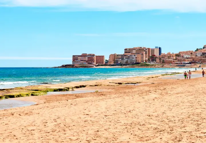 Gente pasea por la **playa de la Mata** con edificios residenciales al fondo y el mar azul.
