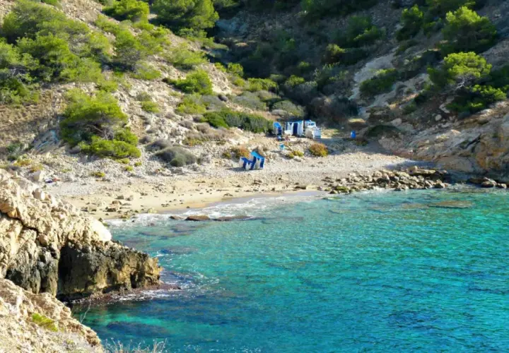 Agua turquesa en la cala Almadrava de Benidorm, con rocas y vegetación en la ladera. Hay algunas sillas y sombrillas en la playa.