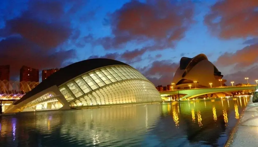 La **Ciudad de las Artes y las Ciencias** de Valencia, con sus icónicos edificios futuristas, se refleja en el agua al anochecer, bajo un cielo dramático.