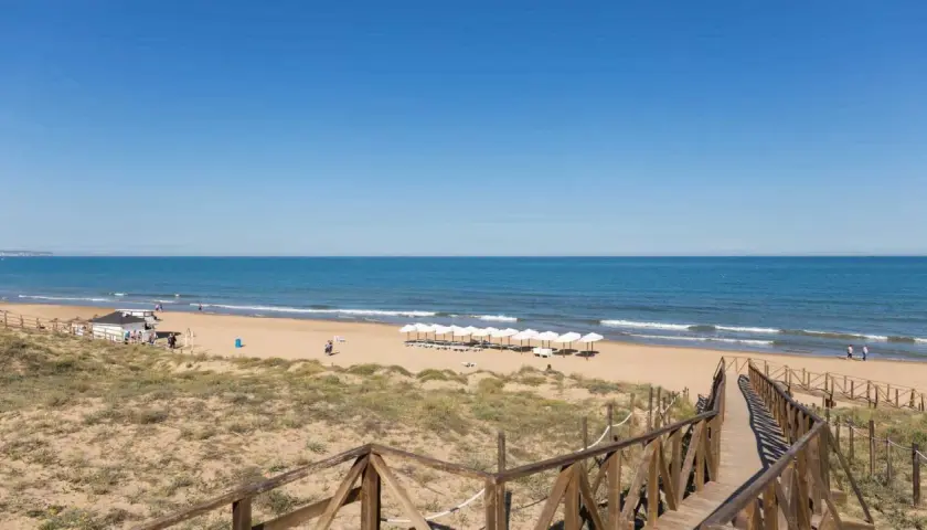 Pasarela de madera hacia la playa de Miramar, con sombrillas blancas alineadas y el mar azul bajo un cielo despejado.