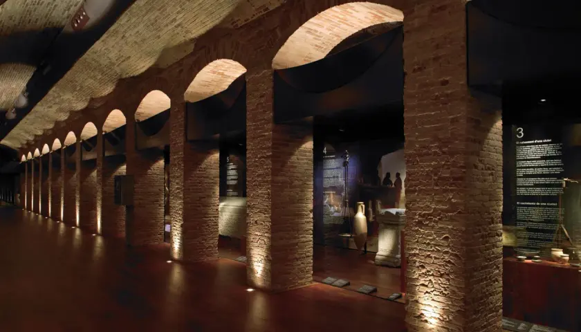 Pasillo del museo de historia de Valencia, con arcos de ladrillo, iluminación tenue y vitrinas con artefactos antiguos.