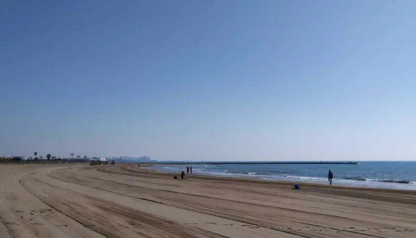 Amplia y tranquila playa de Meliana con arena marcada por vehículos. El mar azul se extiende hasta el horizonte, con algunas personas paseando.