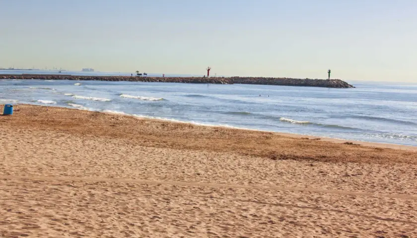 Arena dorada en la playa Port Saplaya, con el mar azul y un espigón con faros.