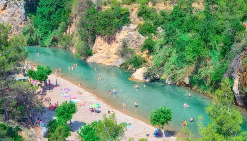 Gente disfrutando del sol y el agua cristalina en el balneario de Montanejos, rodeado de exuberante vegetación y rocas.