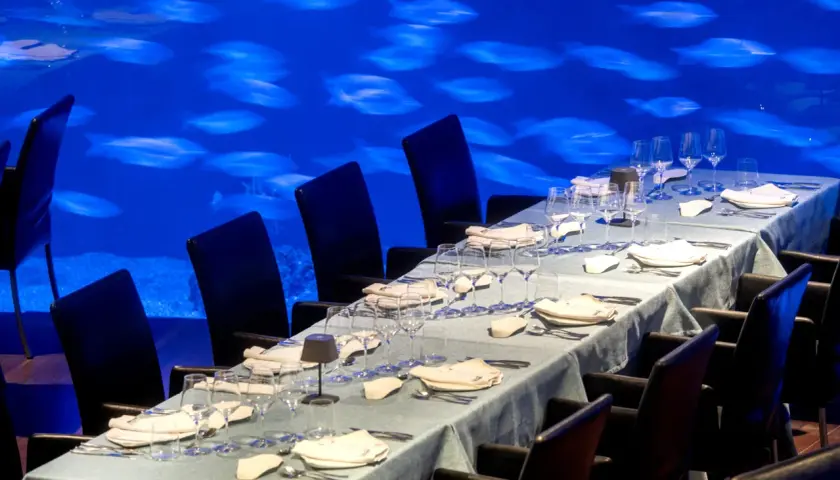 Mesa puesta para cena en el Oceanogràfic, con sillas negras y un acuario de peces de fondo. ¿Dónde comer en el Oceanogràfic? Opiniones.