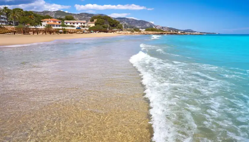 Aguas cristalinas y arena dorada en la playa la Romana, Alcossebre. Un paraíso mediterráneo con montañas al fondo.