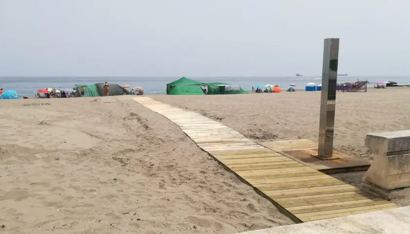 Pasarela de madera hacia las playas con duchas en Alicante. Gente disfrutando del sol bajo sombrillas y tiendas verdes.