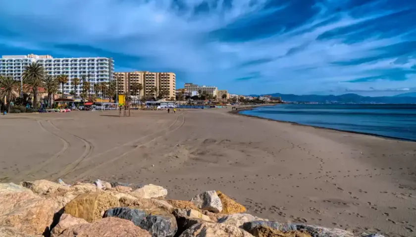 Amplia playa de arena con edificios hoteleros al fondo. El mar azul y el cielo nublado crean un paisaje sereno, ideal para pasear y sentir la **playa fuente de la salud**.