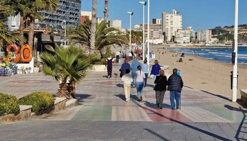 Gente paseando por el **paseo marítimo de Calpe**, con palmeras, edificios altos y la playa al fondo.