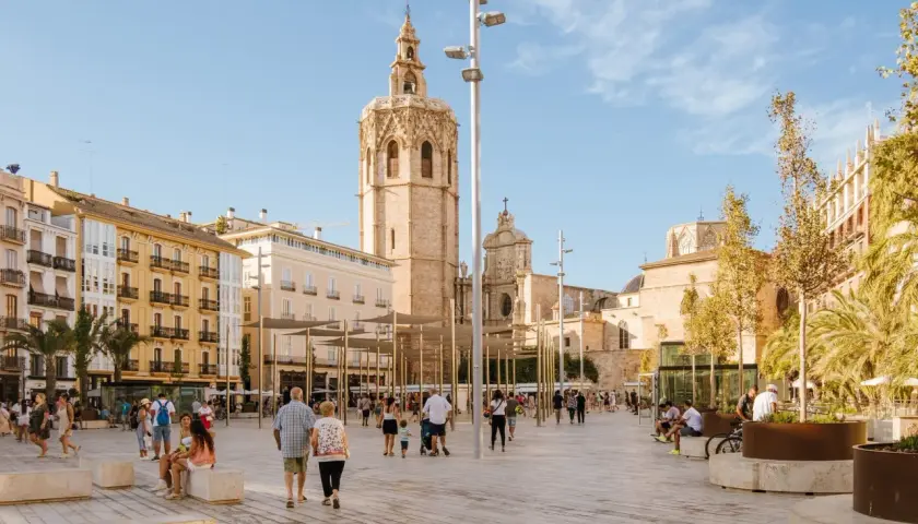 Gente pasea por la plaza frente a la Catedral de Valencia, con el icónico campanario del Miguelete dominando el cielo azul.