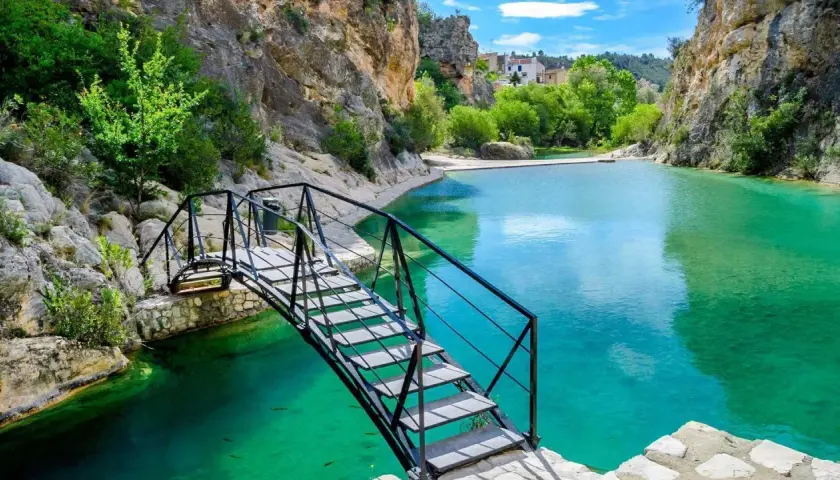 Puente metálico sobre las aguas turquesas del **río Bolbaite**, rodeado de rocas y vegetación exuberante.