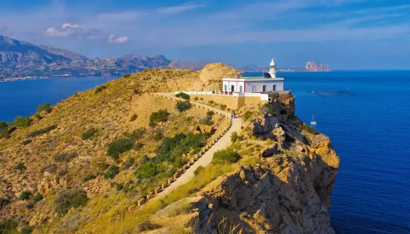 Faro blanco en acantilado rocoso del parque natural de la Serra Gelada. Camino serpenteante, mar azul y montañas al fondo.