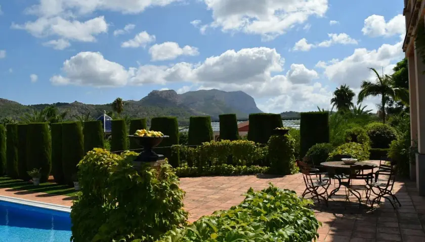 El jardín de l'albarda, con piscina azul, setos verdes, mesa y sillas, y montañas al fondo bajo un cielo nublado.