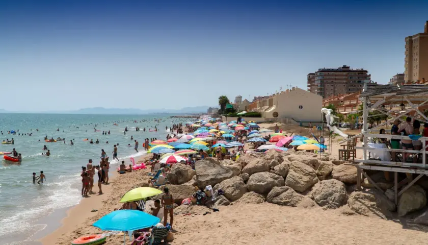 Día soleado en la playa Tavernes de la Valldigna, con mucha gente disfrutando del mar y la arena bajo coloridas sombrillas.