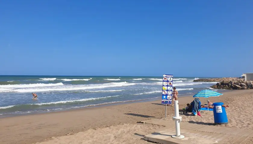 Un día soleado en la playa el Marenyet. Olas rompen en la orilla mientras una persona se baña. Hay un cartel de normas y una sombrilla.