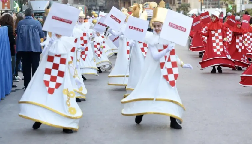 Participantes en la cabalgata del ninot, con trajes blancos y rojos, portan carteles con palabras como "Ciencia" y "Educació".