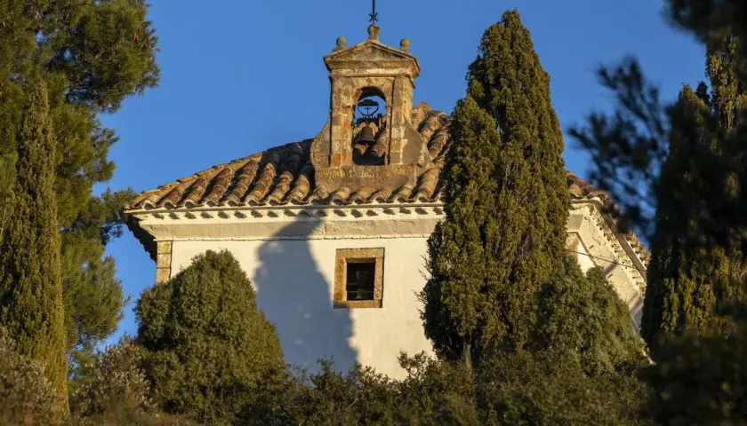 Campanario de piedra con campana, parte del castillo de Villamalefa, rodeado de vegetación y bajo un cielo azul.