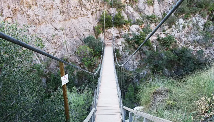 Puentes colgantes de Chulilla, un camino de madera suspendido sobre un barranco rocoso, rodeado de vegetación.