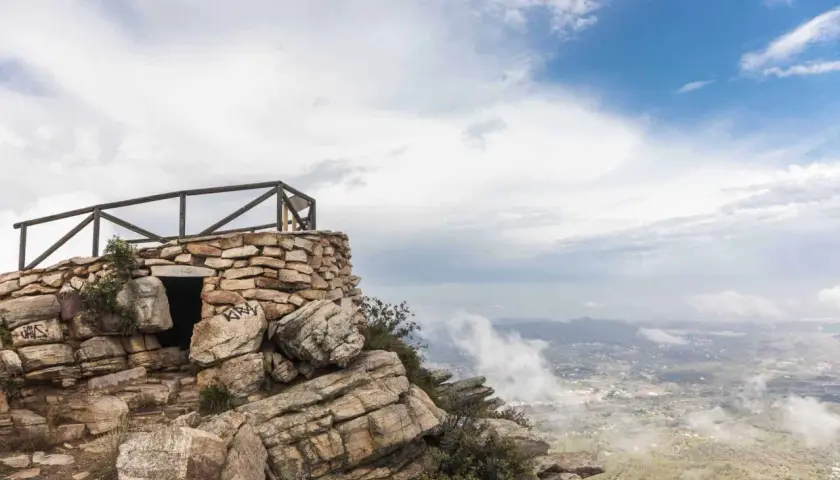 Pequeña construcción de piedra en la cima de una montaña, con barandilla de madera y vistas al valle. El garbí sopla suavemente.