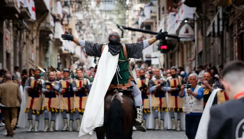 Un jinete con capa blanca lidera un desfile de moros y cristianos, con gente observando y banderas en las calles.