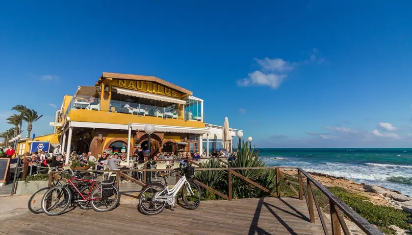 Restaurante Nautilus en la playa Punta Prima, con gente disfrutando en terrazas y bicicletas aparcadas.