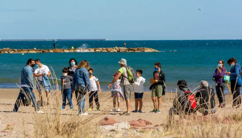 Familias disfrutan de un día soleado en la playa del Puig, con niños jugando y adultos conversando cerca del mar.
