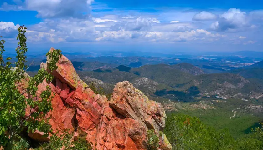 Rocas rojizas y vegetación verde en primer plano, con un vasto paisaje montañoso de la sierra de Espadán bajo un cielo azul con nubes.