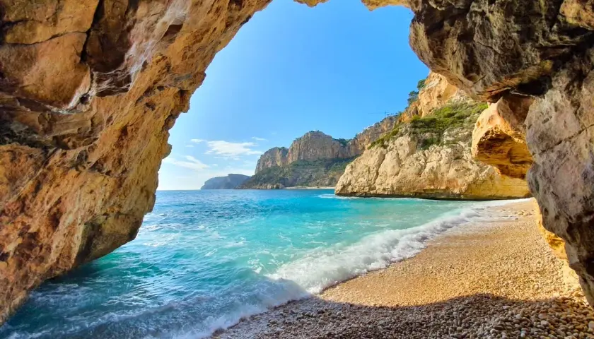 Vista desde una cueva hacia la cala Testos en Benitachell. Aguas turquesas, arena de guijarros y acantilados dorados bajo un cielo azul.