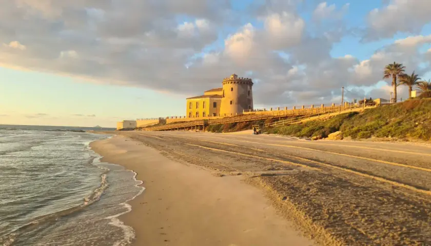 Atardecer en la playa del Conde, con una torre antigua y un paseo de madera. El mar acaricia la arena dorada bajo un cielo nublado.