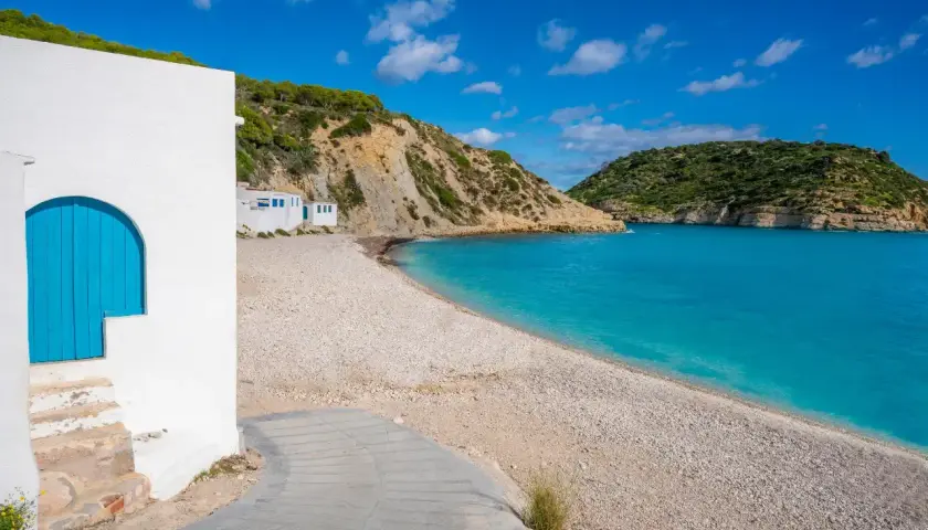 Casa blanca con puerta azul en la playa de guijarros de Vall d'Ebo, con aguas turquesas y colinas verdes.