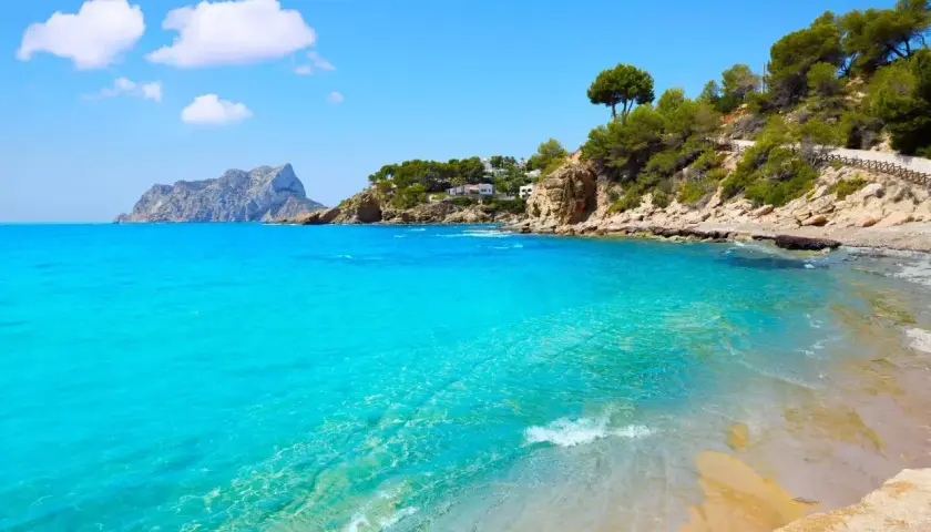 Aguas turquesas y arena dorada en la cala Pinets, con rocas y pinos coronando la costa. Un paraíso mediterráneo.
