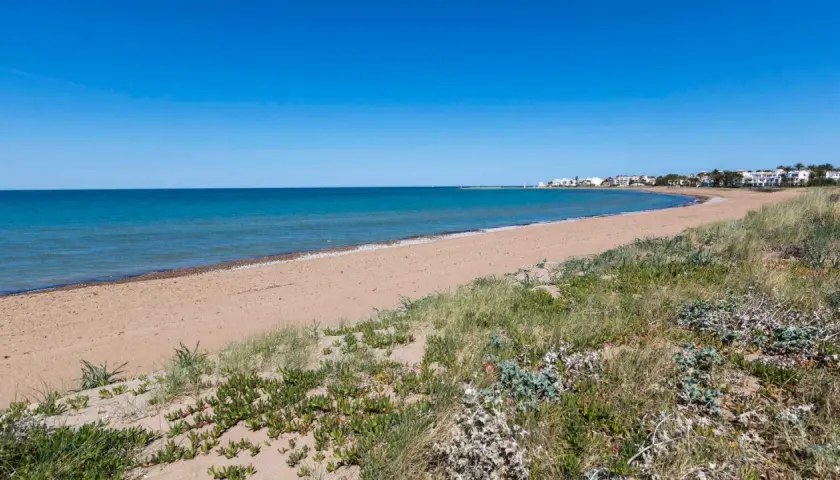 La playa Els Molins se extiende con arena dorada y vegetación costera bajo un cielo azul. Al fondo, se vislumbran casas blancas y un faro.