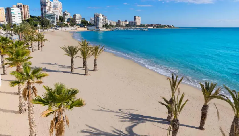 La **playa de Altea** se extiende con arena dorada y palmeras. El mar Mediterráneo, de un azul turquesa intenso, baña la costa, mientras edificios modernos se alzan al fondo.