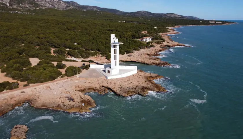 Faro blanco moderno en la costa rocosa de la Sierra de Irta, con el mar azul y montañas verdes al fondo.