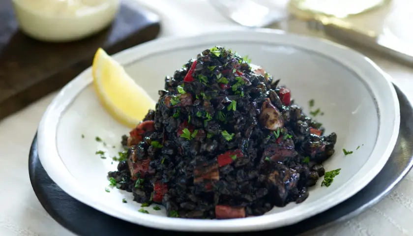 Delicioso arroz negro con trozos de calamar y pimiento rojo, adornado con perejil fresco y una rodaja de limón.