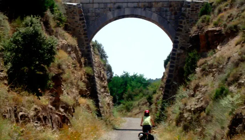 Ciclista pedalea por la vía verde Valencia bajo un antiguo puente de piedra.