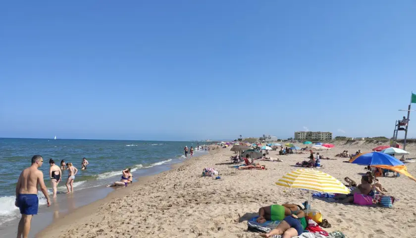 Gente disfrutando del sol y el mar en la playa de la Garrofera. Sombrillas, toallas y bañistas llenan la arena dorada bajo un cielo azul.