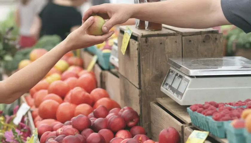 Manos intercambian una manzana en un mercadillo de oliva. Tomates, frambuesas y melocotones esperan ser pesados.