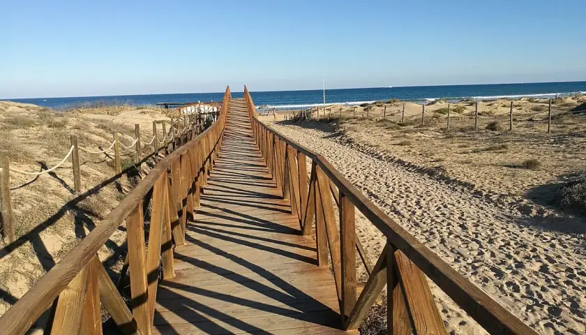 Pasarela de madera que conduce a la playa del campo, con dunas y el mar azul al fondo.