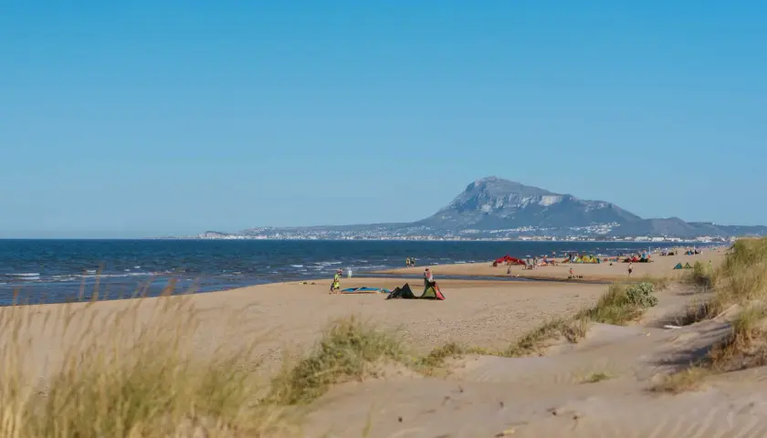 Dunas y mar en la playa de Rabdells, con gente disfrutando del sol y el paisaje montañoso al fondo.
